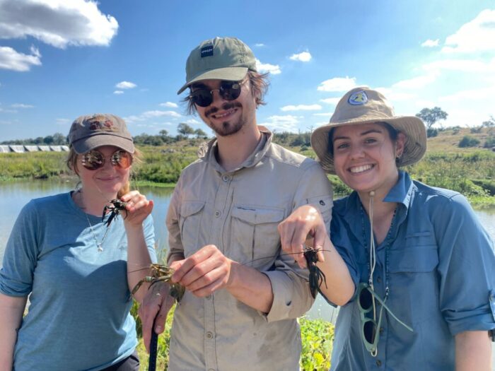 Three young postgraduate researchers standing in sunshine beside a river, holding crayfish.